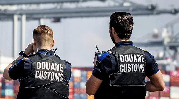 Two customs officers wearing "DOUANE CUSTOMS" vests at a shipping port, facilitating Landmark Global logistics.