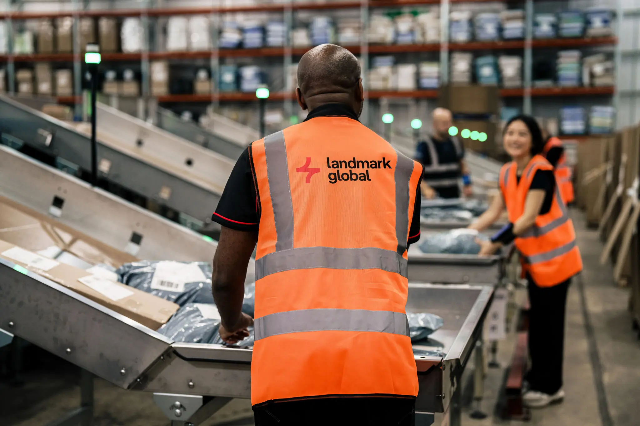 Warehouse workers in orange high-visibility vests with the Landmark Global logo sorting parcels on a conveyor belt.